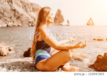 Yoga on the beach. A happy woman meditating in a yoga pose on the beach, surrounded by the ocean and rock mountains, promoting a healthy lifestyle outdoors in nature, and inspiring fitness concept. 120941905