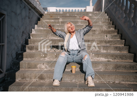 A woman in jeans and a jacket sits on a set of stairs. She is smiling and holding a cup. A woman in jeans and a jacket sits on a set of stairs. She is smiling and holding a cup. 120941986
