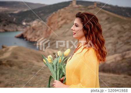 Portrait of a happy woman with long hair against a background of mountains and sea. Holding a bouquet of yellow tulips in her hands, wearing a yellow sweater Portrait of a happy woman with long hair against a background of mountains and sea. Holding a bouquet of yellow tulips in her hands, wearing a yellow sweater 120941988