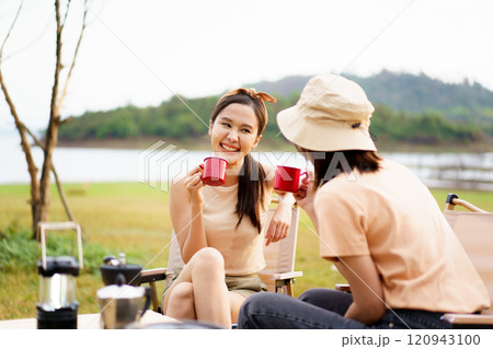 Happy cheerful Asian young women - friends camping in the lake forest and enjoy drinking a cup of tea or coffee. Female friends on a camping trip in the natural park in summer. 120943100