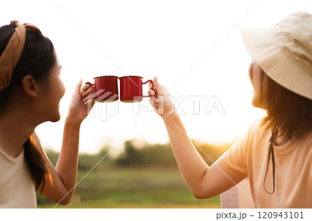 Happy cheerful Asian young women - friends camping in the lake forest and enjoy drinking a cup of tea or coffee. Female friends on a camping trip in the natural park in summer. 120943101