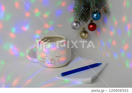 A Christmas branch with balloons hangs above the table, a spiral notebook with a blue pen and a white cup in the shape of a cat's head are on the table A Christmas branch with balloons hangs above the table, a spiral notebook with a blue pen and a white cup in the shape of a cat's head are on the table 120956501