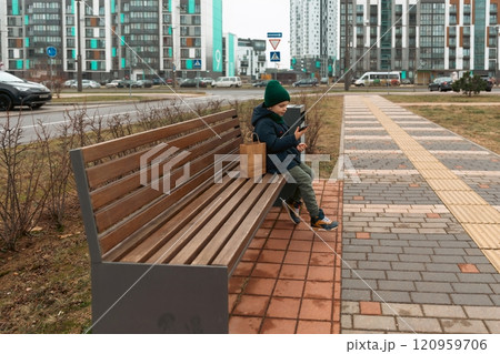 Young boy sitting on a park bench with a smartphone in a modern urban area during a cloudy day 120959706