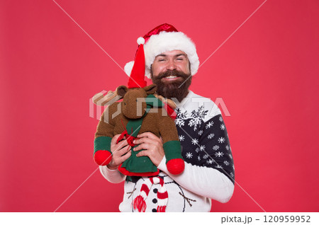 Christmas prep concept. Hipster bearded face winter sweater. Kindness and charity. Winter holiday. Buy gifts. Winter sale. Winter feast. Reindeer toy. Man santa hat hold christmas gift red background 120959952