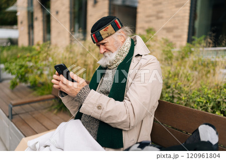 Focused aged homeless man sitting on city bench, using smartphone, trying to connect with family or access essential services. Concept of poverty and vulnerability, evoking empathy and compassion. 120961804