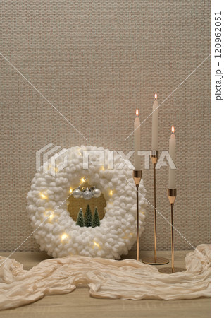 Christmas decor, white soft plush wreath with lights, three lit candles in golden holder, beige cloth on wooden table 120962051