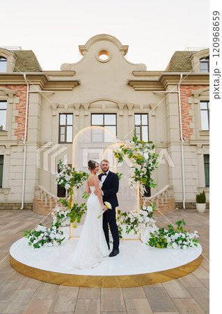 The bride and groom on a wedding arch decorated with greenery and white flowers. 120968659