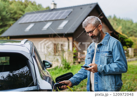 Man charging electric car in front of his house, plugging the charger into the charging port. House with solar panel system on roof behind him. 120972048