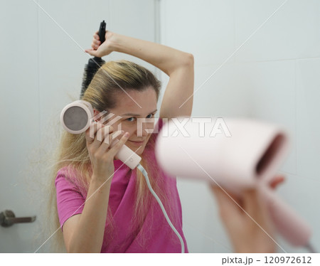 A woman is expertly styling her hair using a blow dryer and brush while in the bathroom A woman is expertly styling her hair using a blow dryer and brush while in the bathroom 120972612
