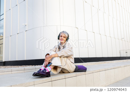 Smiling Trendy Mature Woman Listening To Music On Headphones While Sitting On Steps Of White Modern Building In City 120974043