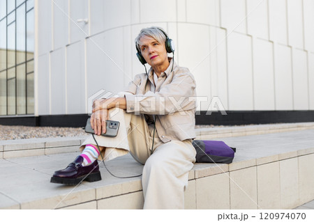 Beautiful Trendy Senior Woman with Short Hair and Headphones in Beige Clothing Sitting Outdoors in City, Listening to Music, Podcast or Audiobook Against Modern White Building 120974070