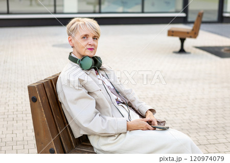 Elegant Elderly Woman with Short Hair Wearing Headphones and Using Smartphone While Sitting on Bench Outdoors in City, Active lifestyle for Older adults Elegant Elderly Woman with Short Hair Wearing Headphones and Using Smartphone While Sitting on Bench Outdoors in City, Active lifestyle for Older adults 120974079
