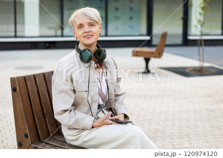Beautiful Smiling Mature Woman with Short Hair Wearing Headphones and Using phone While Sitting on Bench Outdoors in City, Active lifestyle for Older adults 120974120