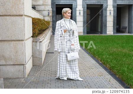 Fashionable Mature Woman with Short Hair Standing Outside in City Dressed in Stylish Checkered White Suit with Blazer and Trousers, Accessories and Handbag Showcasing Trendy Clothing and Street Style 120974135