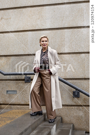 Stylish Woman Wearing Fashionable Clothing Beige Coat and trousers, Felt Hat, Accessories and Leather Gloves Walking Stairs Against Backdrop of Urban Buildings in City, Street Style and Fashion Trends 120974154