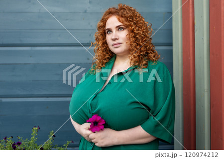 Plus Size Woman with Curly Red Hair in Green Clothing Holds Flower Against Vibrant Blue Wall Background, Beauty and Confidence Outdoors 120974212