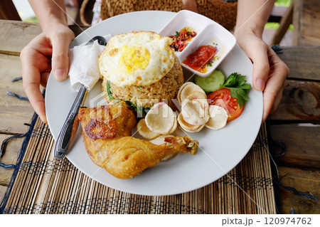 Woman hand holding Pecel ayam or penyet ayam is fried chicken with rice and fried eggs decorated with tomato and spicy sauce is tradition food of Indonesia Woman hand holding Pecel ayam or penyet ayam is fried chicken with rice and fried eggs decorated with tomato and spicy sauce is tradition food of Indonesia 120974762