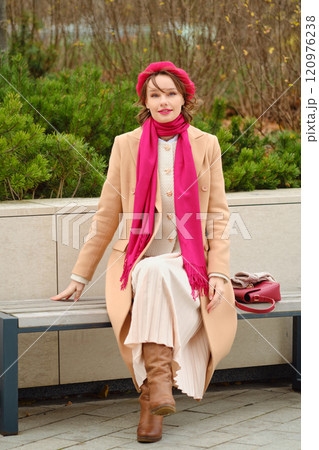 A woman in a fashionable coat and scarf relaxing on a bench in a park during autumn 120976238