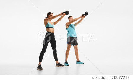 Full-length image of young sportive man and woman with sportive bodies, training, doing exercises with kettlebell against white studio background Full-length image of young sportive man and woman with sportive bodies, training, doing exercises with kettlebell against white studio background 120977218