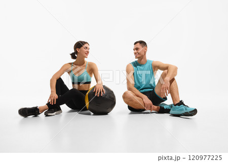 Young slim woman and muscular man, couple in sportswear sitting on floor and preparing for training with medicine ball against white studio background Young slim woman and muscular man, couple in sportswear sitting on floor and preparing for training with medicine ball against white studio background 120977225