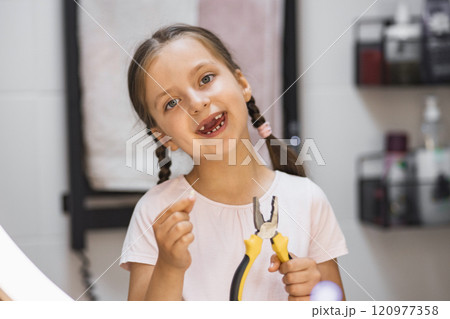Caucasian girl smiling in bathroom holding fallen baby tooth and pliers, reflecting excitement and achievement after tooth loss. 120977358