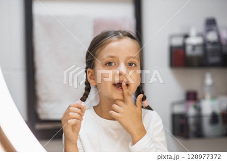 Caucasian girl with pigtails holding fallen baby tooth, looking into mirror in bathroom, capturing childhood milestone moment. 120977372