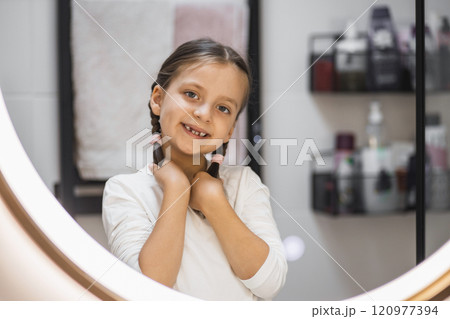 Little Caucasian girl with braided hair smiling at mirror, showing missing baby teeth. Reflective and joyful moment captured in a modern bathroom setting. 120977394