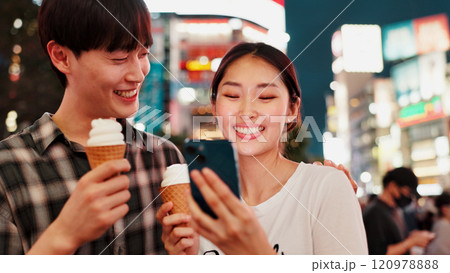 Couple, ice cream and happy with phone for travel, date or notification with bonding, love or contact in city. People, man and woman on smartphone, laugh or gelato on social network at night in Japan Couple, ice cream and happy with phone for travel, date or notification with bonding, love or contact in city. People, man and woman on smartphone, laugh or gelato on social network at night in Japan 120978888