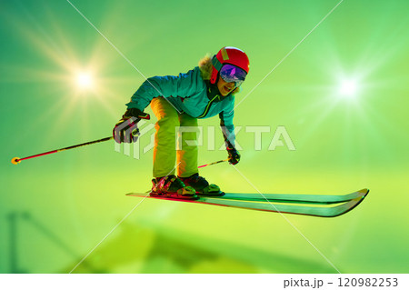 Low angle, side view of female athlete, skier in colorful gear mid-air jump neon green background. Dynamic pose. 120982253