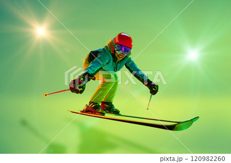 Low angle, side view of female athlete, skier in colorful gear mid-air jump neon green background. Dynamic pose. Low angle, side view of female athlete, skier in colorful gear mid-air jump neon green background. Dynamic pose. 120982260