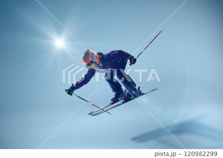 Mid-air photo of skier in vibrant winter attire strikes a streamlined position. Her skis hover parallel as they glide against clean blue background. 120982299