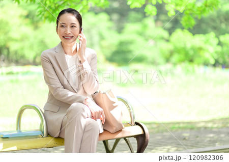 公園のベンチに座りながらスマートフォンで会話するビジネスウーマン 120982306