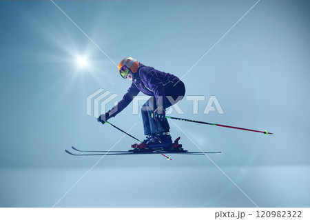 Side view of athlete executing precise ski jump, wearing purple jacket and mirrored goggles against blue backdrop and sunlight enhancing action Side view of athlete executing precise ski jump, wearing purple jacket and mirrored goggles against blue backdrop and sunlight enhancing action 120982322
