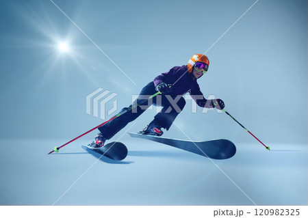 Winter athlete launching dynamically, orange helmet and reflective goggles glinting under studio lighting, emphasizing speed against seamless blue. 120982325