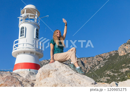 Woman sitting on rocks near a white and red lighthouse, waving under a clear blue sky. Capturing family vacation, freedom, coastal exploration, perfect for travel and maritime themes. 120985275