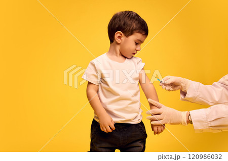 Boy, child in white t-shirt looking cautiously at syringe held by doctor during vaccination session against yellow studio background. Boy, child in white t-shirt looking cautiously at syringe held by doctor during vaccination session against yellow studio background. 120986032