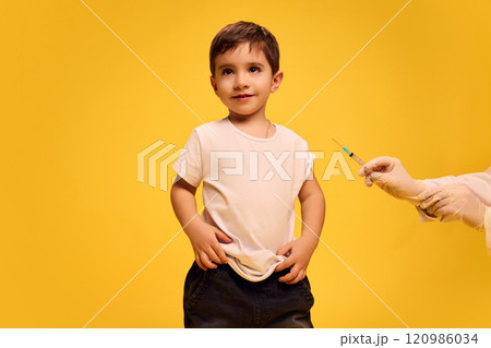 Little boy, child in black and white clothes calmly standing while doctor making vaccine injection with syringe against yellow studio background. Little boy, child in black and white clothes calmly standing while doctor making vaccine injection with syringe against yellow studio background. 120986034