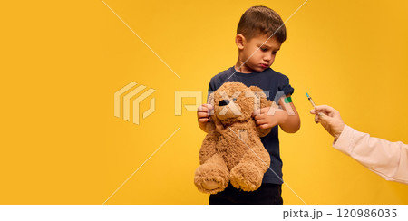 Sad 5 year old boy, child holding teddy bear toy while doctor holding syringe and making vaccination against yellow studio background. Sad 5 year old boy, child holding teddy bear toy while doctor holding syringe and making vaccination against yellow studio background. 120986035