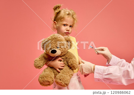 Little sad girl, child holding teddy bear toy while doctor holding syringe and making vaccination against pink studio background Little sad girl, child holding teddy bear toy while doctor holding syringe and making vaccination against pink studio background 120986062