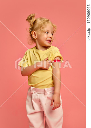 Smiling little girl, child in yellow t-shirt, pointing on her arm with red plaster after vaccination injection, against pink studio background Smiling little girl, child in yellow t-shirt, pointing on her arm with red plaster after vaccination injection, against pink studio background 120986068