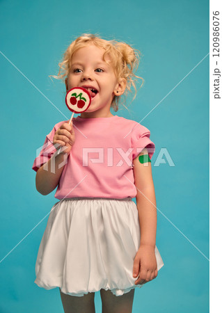 Happy little child, girl with green plaster on arm after vaccination, licking lollipop against blue studio background. Brave child. Happy little child, girl with green plaster on arm after vaccination, licking lollipop against blue studio background. Brave child. 120986076