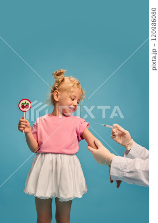 Smiling and curious little girl in pink t-shirt and white skirt, holding lollipop and looking how doctor making vaccination against blue studio background Smiling and curious little girl in pink t-shirt and white skirt, holding lollipop and looking how doctor making vaccination against blue studio background 120986080