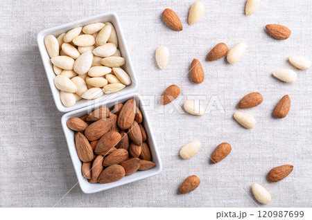Almonds in square shaped white bowls, on linen fabric. Almonds with seed coats and blanched. Fruits of Prunus amygdalus, syn. Prunus dulcis, ready-to-eat as snack or as ingredient for baking. Photo. 120987699