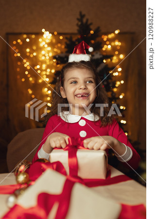 Happy laughing 6 years old girl in Santa Claus costume wraps gifts boxes with red ribbons and festive lights on background Happy laughing 6 years old girl in Santa Claus costume wraps gifts boxes with red ribbons and festive lights on background 120988227