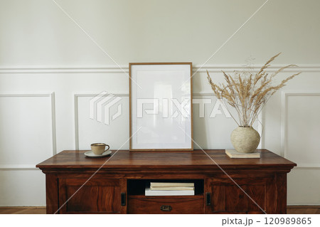 Scandinavian interior still life. Modern living room. Blank picture mock up poster frame. Mahogany wooden dresser sideboard. Vase with dry flowers, books Cup of tea, coffee, white wall background. 120989865