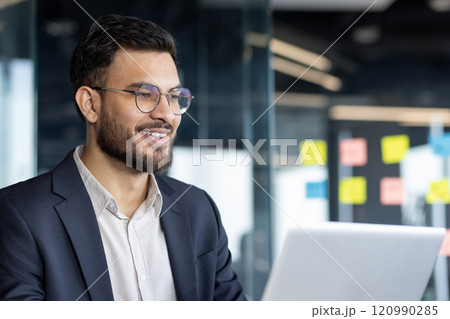 A confident businessman is smiling as he works on his laptop in a bright office setting. His expression conveys professionalism and focus in a modern and productive corporate environment. 120990285