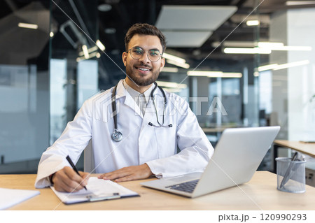 A professional doctor with a stethoscope sits at a desk, smiling confidently while working on paperwork and using a laptop in an office setting. A professional doctor with a stethoscope sits at a desk, smiling confidently while working on paperwork and using a laptop in an office setting. 120990293