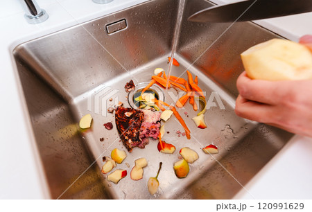 Peeling potatoes over a kitchen sink filled with food scraps and running water. 120991629