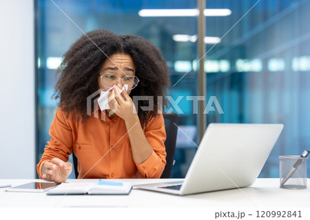 A young woman sneezes into a tissue while working at her desk with a laptop. Her expression shows discomfort, highlighting workplace health and productivity challenges. 120992841