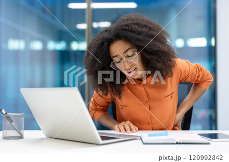Young businesswoman feeling discomfort due to back pain at her desk. She leans forward while working on a laptop, highlighting workplace ergonomics challenges. Stress and discomfort visible. Young businesswoman feeling discomfort due to back pain at her desk. She leans forward while working on a laptop, highlighting workplace ergonomics challenges. Stress and discomfort visible. 120992842